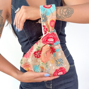 Peut inclure: Un sac poignet en tissu beige orné de broderies florales aux tons vifs de rouge, rose, orange et turquoise. Le sac présente une anse nouée et une matière de base transparente, mettant en valeur un motif dense de fleurs et de feuilles.