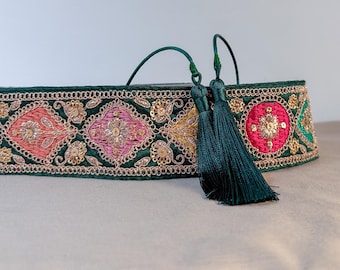 Può includere: Fascia decorativa ricamata con intricati motivi floreali rosa, oro e rossi su base verde scuro. Presenta paillettes dorate e un bordo dettagliato. Due nappe verde scuro pendono da un lato. Larghezza di circa 5 cm.
