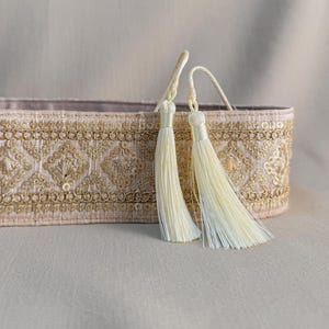 Pode incluir: Caixa decorativa retangular rosa pálido com bordados e lantejoulas douradas. Duas borlas creme pendem do topo. O padrão é detalhado e ornamentado, adequado para guardar ou exibir.