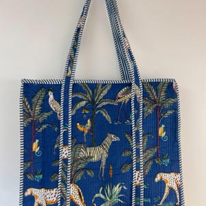 May include: A blue tote bag with a jungle-themed print featuring zebras, leopards, birds, and palm trees. The bag has striped handles and trim, and measures approximately 15 inches wide and 14 inches tall.