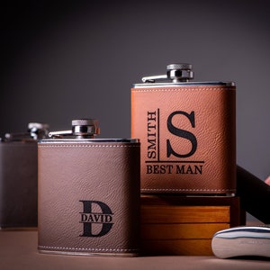 Könnte beinhalten: Zwei personalisierte, lederbezogene Edelstahl-Taschenflaschen. Eine Flasche ist braun mit dem Text "SMITH BEST MAN" und dem Initial "S". Die andere Flasche ist grau mit dem Text "DAVID" und dem Initial "D".