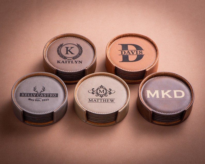 May include: Set of six brown leather coasters with different personalized engravings. The coasters are stacked in a holder. The engravings include "Kaitlyn" with a "K" initial, "Davis" with a "D" initial, "Kelly Castro" with a deer antler design, "Matthew" with an "M" initial, and "MKD".