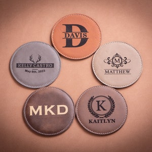 May include: Five personalized leather coasters with different designs. The coasters are brown, gray, and beige. The designs include a monogram, a deer antler, a floral design, and a wreath. The text on the coasters reads "Kelly Castro May 6th, 2023", "Davis", "Matthew", "MKD", and "Kaitlyn".