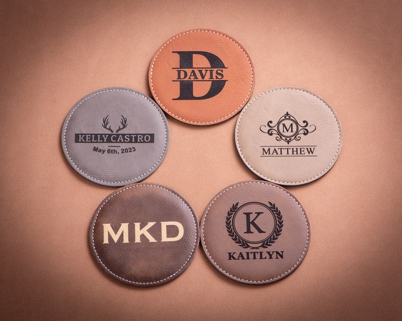 May include: Five personalized leather coasters with different designs. The coasters are brown, gray, and beige. The designs include a monogram, a deer antler, a floral design, and a wreath. The text on the coasters reads "Kelly Castro May 6th, 2023", "Davis", "Matthew", "MKD", and "Kaitlyn".