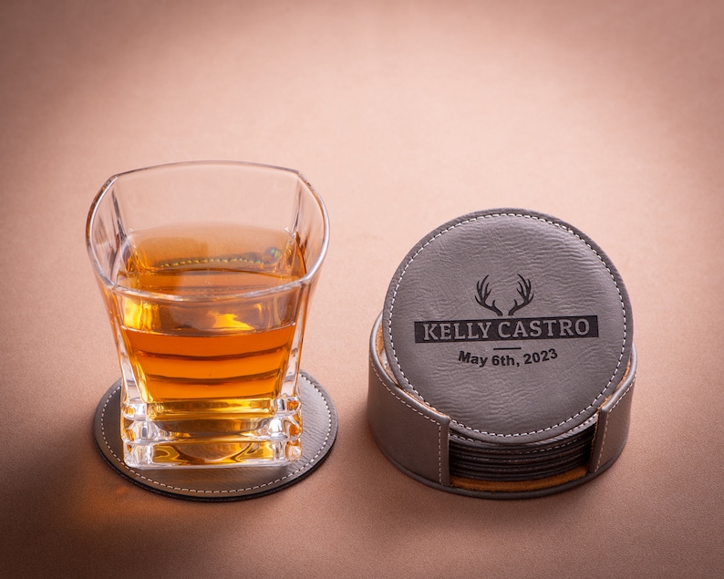 May include: A set of six gray leather coasters with a brown leather base. The top coaster features a personalized inscription with the text "KELLY CASTRO May 6th, 2023" and a stylized deer antler design.