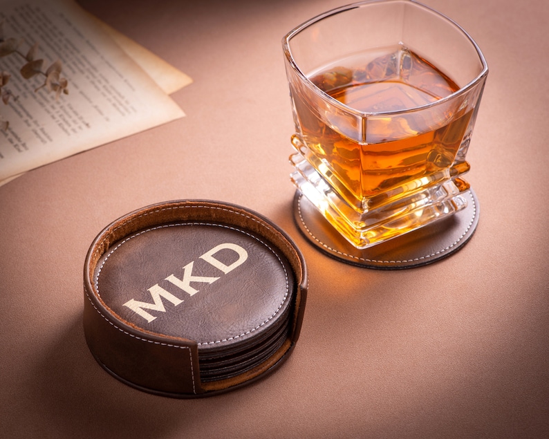 May include: A set of six brown leather coasters with a stitched edge. The top coaster has the initials "MKD" embossed in gold lettering. A glass of amber liquid sits on a brown leather coaster.