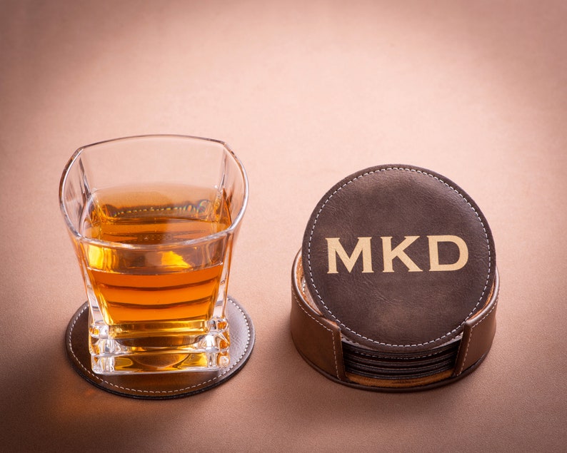 May include: A set of brown leather coasters with a holder. The top coaster has the initials "MKD" in gold lettering.