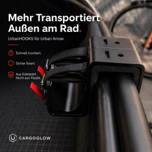 May include: A black UrbanHOOKS cargo carrier for a bicycle, designed for the Urban Arrow model. The carrier is mounted on a black bar and secured with black straps and red accents. The text on the image says "Mehr Transportiert Außen am Rad."