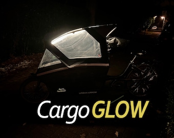 Puede incluir: Una bicicleta de carga negra con una cubierta de plástico transparente y un texto blanco "Cargo GLOW" en la parte delantera. La bicicleta está aparcada de noche y la cubierta está iluminada desde el interior.