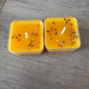 May include: Two square candles with a bright yellow wax and a white wick. The candles are in clear plastic containers and are decorated with dried flowers. The candles are placed on a gray wooden surface.