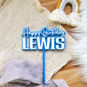 May include: Blue and white cake topper with the text "Happy Birthday Lewis" in a cursive font.