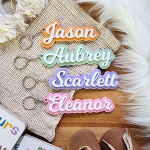 May include: Four colourful acrylic keychains with the names "Jason", "Aubrey", "Scarlett", and "Eleanor" written in white. The keychains are on a beige fabric surface.