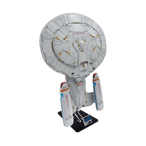 May include: A model of the Star Trek USS Enterprise NCC-1701-D, made from building blocks. The model is mainly white with grey and yellow details, and has two blue and red engine nacelles. It is displayed on a black stand.