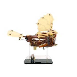 Pode incluir: Um modelo LEGO de uma máquina voadora em marrom e dourado com um suporte de exibição de acrílico transparente. O modelo tem uma vela grande de cor bege e uma pequena figura de um mago.
