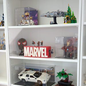 Può includere: Una libreria bianca espone vari oggetti da collezione all'interno di teche trasparenti. Gli articoli includono un logo Marvel, una maschera di Spider-Man, una nave Lego Star Wars e un modello di auto sportiva bianca. Le mensole sono contro un muro bianco.