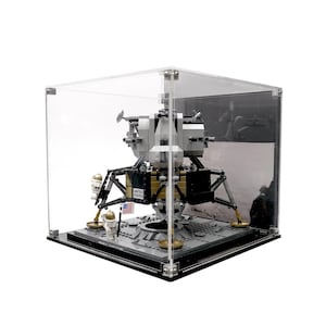 May include: A Lego model of the Apollo 11 lunar lander, displayed in a clear acrylic case. The model is grey and gold, with the words "UNITED STATES" visible. Includes two astronaut figures and a small American flag.