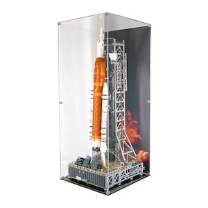 Könnte beinhalten: Detailliertes Modell einer Rakete mit orangefarbenen und weißen Abschnitten, präsentiert in einer transparenten Acrylvitrine. Das Modell steht auf einer Startplattform mit detaillierter Tragstruktur, vor dem Hintergrund einer feurigen Weltraumszene.