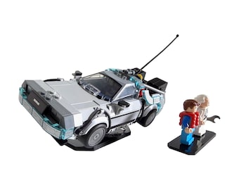 May include: A silver DeLorean time machine model with black accents and blue lights, along with miniature figures of Marty McFly and Doc Brown. The model has a black antenna and is set against a white background.
