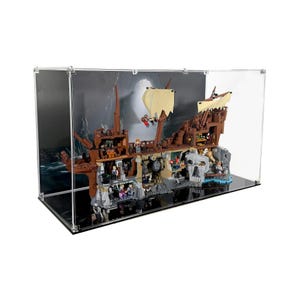 Könnte beinhalten: Ein detailliertes braunes Lego-Piraten-Schiffsmodell mit beigen Segeln, ausgestellt in einem transparenten Acrylgehäuse. Das Schiff ist vor dem Hintergrund eines stürmischen Meeres und einer schädelartigen Felsformation platziert.