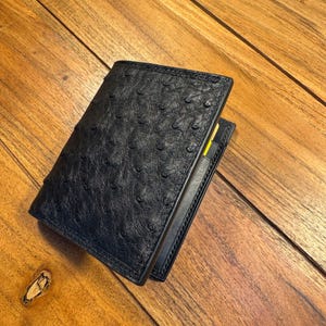 Può includere: Un portafoglio nero a due ante caratterizzato da una texture in pelle di struzzo con un design distintivo a follicoli di piuma. Il portafoglio è parzialmente aperto, rivelando il bordo di una carta gialla inserita nello scomparto interno.