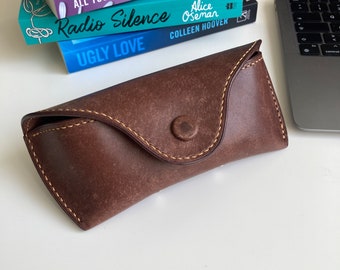 May include: A brown leather glasses case with a button closure and yellow stitching.