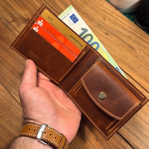 Könnte beinhalten: Ein braunes Leder-Portemonnaie mit mehreren Kartenfächern, einem Münzfach mit goldenem Druckknopf und einem Geldscheinfach, das einen 100-Euro-Schein enthält. Das Portemonnaie weist sichtbare Nähte und eine Prägung auf.