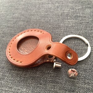 May include: A brown leather key chain with a round, stitched pocket and a metal ring. The key chain is lying on a gray fabric surface.