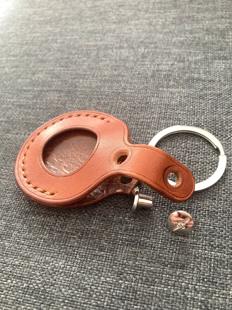May include: A brown leather key chain with a round, stitched pocket and a metal ring. The key chain is lying on a gray fabric surface.