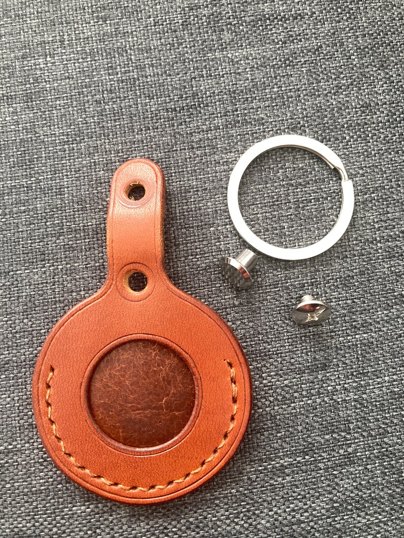 May include: A brown leather key fob with a stitched circle in the center and a metal ring attached. The key fob has two holes for attaching to a key ring.