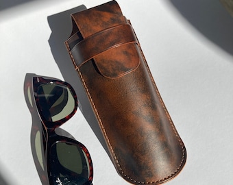 May include: A brown leather sunglasses case with a flap closure and stitching detail. A pair of tortoiseshell Ray-Ban sunglasses are lying next to the case.