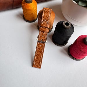 May include: A brown leather watch strap with white stitching and a silver buckle. The strap is textured and lies on a white surface, surrounded by spools of thread in various colors, including orange, black, and red.
