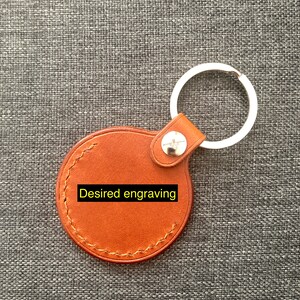 May include: A brown leather keychain with a round tag that says "Desired engraving". The keychain has a silver metal ring.