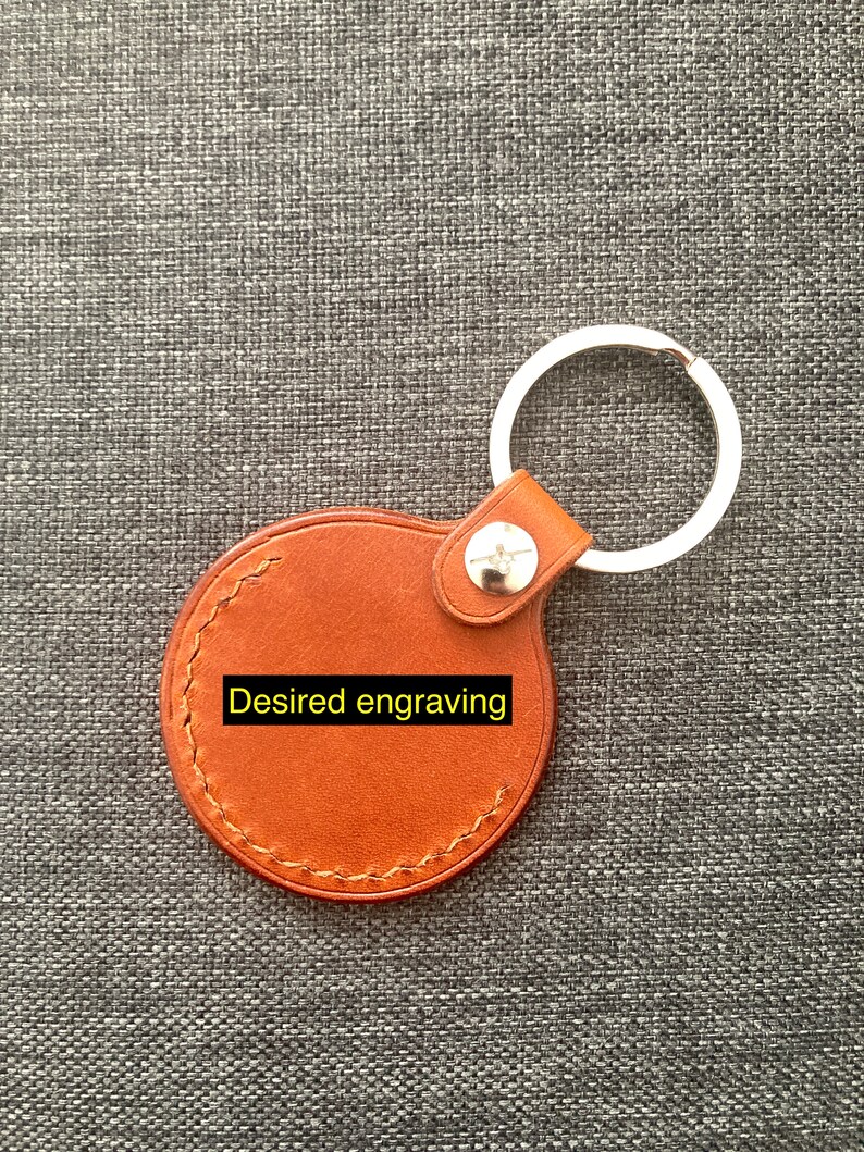 May include: A brown leather keychain with a round tag that says "Desired engraving". The keychain has a silver metal ring.