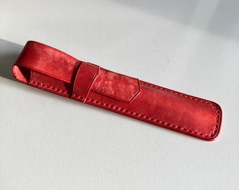 Puede incluir: Un estuche para bolígrafos de cuero rojo con cierre de solapa y detalles de costura.