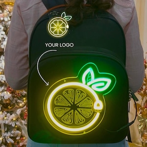 May include: Black backpack with a neon lemon design. The backpack features a large, illuminated lemon slice graphic on the front, with the text "YOUR LOGO" above. The neon design glows in yellow and green.