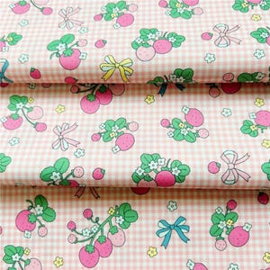 Peut inclure: Tissu rose &agrave; carreaux avec un motif r&eacute;p&eacute;t&eacute; de fraises, de feuilles vertes, de fleurs blanches et de n&oelig;uds roses, jaunes et bleus. Le tissu est pli&eacute;, montrant plusieurs couches du motif.