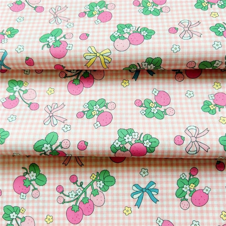 Peut inclure: Tissu rose &agrave; carreaux avec un motif r&eacute;p&eacute;t&eacute; de fraises, de feuilles vertes, de fleurs blanches et de n&oelig;uds roses, jaunes et bleus. Le tissu est pli&eacute;, montrant plusieurs couches du motif.