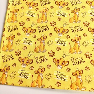 Puede incluir: Tela amarilla con un patr&oacute;n repetido de un cachorro de le&oacute;n de dibujos animados con una corona. El texto "Little King" se repite en todo el patr&oacute;n.