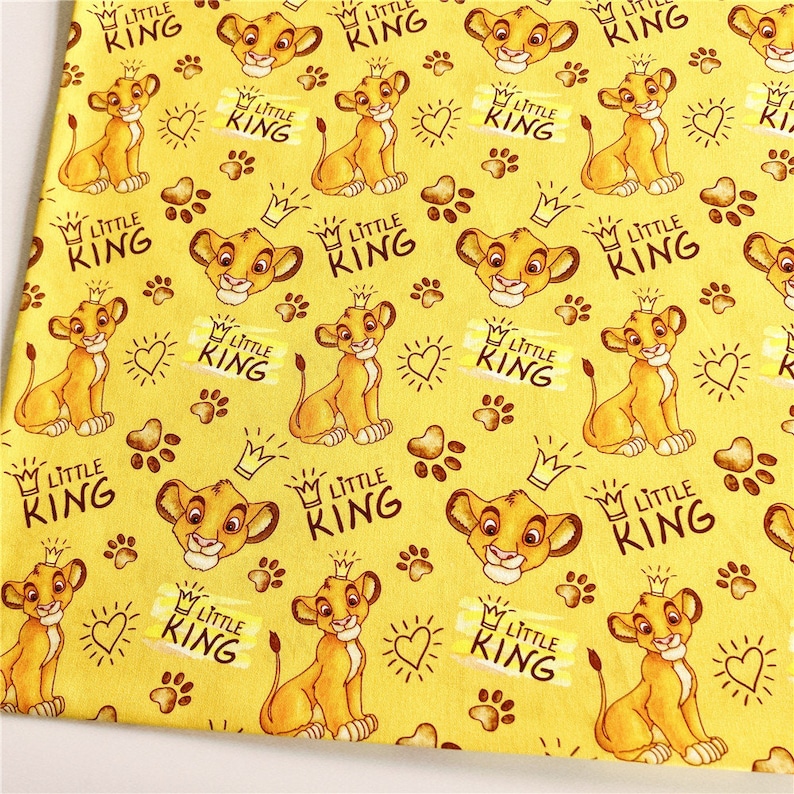 Puede incluir: Tela amarilla con un patr&oacute;n repetido de un cachorro de le&oacute;n de dibujos animados con una corona. El texto "Little King" se repite en todo el patr&oacute;n.