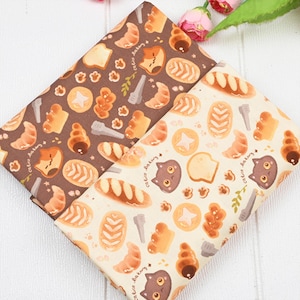 Op de afbeelding: Twee stukken stof met een herhaald patroon van bakkerijproducten en kattengezichten. De ene stof is bruin, de andere is gebroken wit. Het ontwerp bevat croissants, broden en stervormige koekjes. De tekst "calico Bakery" is ook aanwezig.