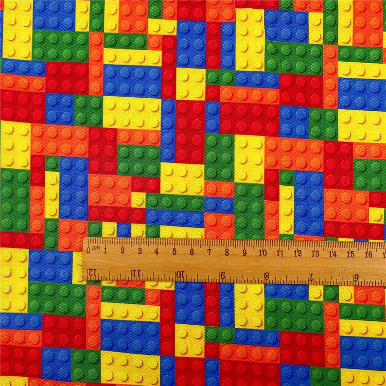 K&ouml;nnte beinhalten: Ein farbenfroher Stoff mit einem sich wiederholenden Muster aus roten, gelben, blauen und gr&uuml;nen Plastikbausteinen. Der Stoff wird mit einem Lineal gemessen, das 16 Zentimeter anzeigt.