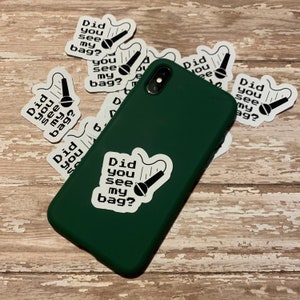 May include: A green phone case with a white sticker that says "Did you see my bag?" and a microphone graphic. The sticker is surrounded by other stickers with the same text and graphic.