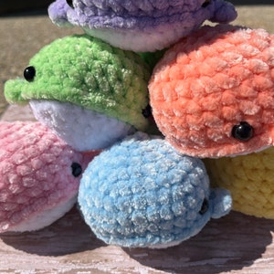 May include: A stack of six crocheted plush whale toys in various pastel colors, including purple, green, orange, pink, blue, and yellow. Each whale has a white belly and black eyes.
