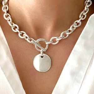 Peut inclure: Un collier argenté avec une chaîne épaisse. Le collier est orné d'un pendentif rond et d'une fermeture à bascule. Le bijou est porté sur une chemise blanche.