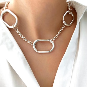 Peut inclure: Un collier en chaîne argentée avec trois gros maillons ovales. Le collier est porté par un mannequin portant une chemise blanche.