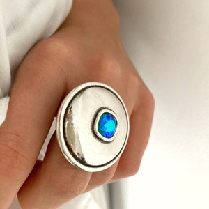 Peut inclure: Une bague argentée de grande taille, de forme circulaire. La bague présente une face proéminente et ronde avec une pierre précieuse centrale bleu-vert de forme carrée. La bague est portée au doigt, sur fond blanc.