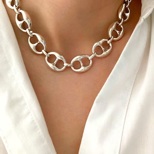 Può includere: Una collana a catena argentata con grandi maglie ovali. La collana è indossata attorno al collo e le maglie hanno un design distintivo con due aperture in ogni ovale. Un gioiello audace e di tendenza.