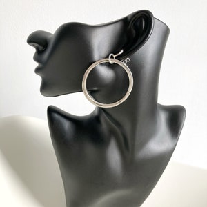 Peut inclure: Boucles d'oreilles créoles argentées avec un design circulaire simple. Les boucles d'oreilles sont présentées sur une tête de mannequin noire.