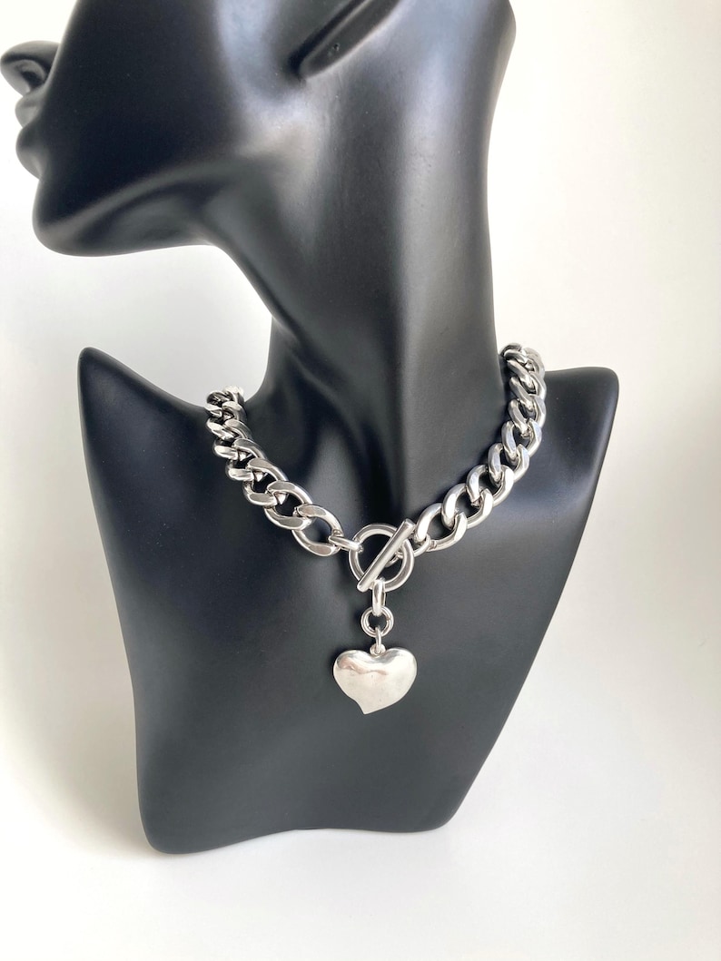 K&ouml;nnte beinhalten: Eine silberne Kettenkette mit einem gro&szlig;en, klobigen Gliederdesign. Die Kette hat einen Toggle-Verschluss und einen baumelnden Herz-Anh&auml;nger.