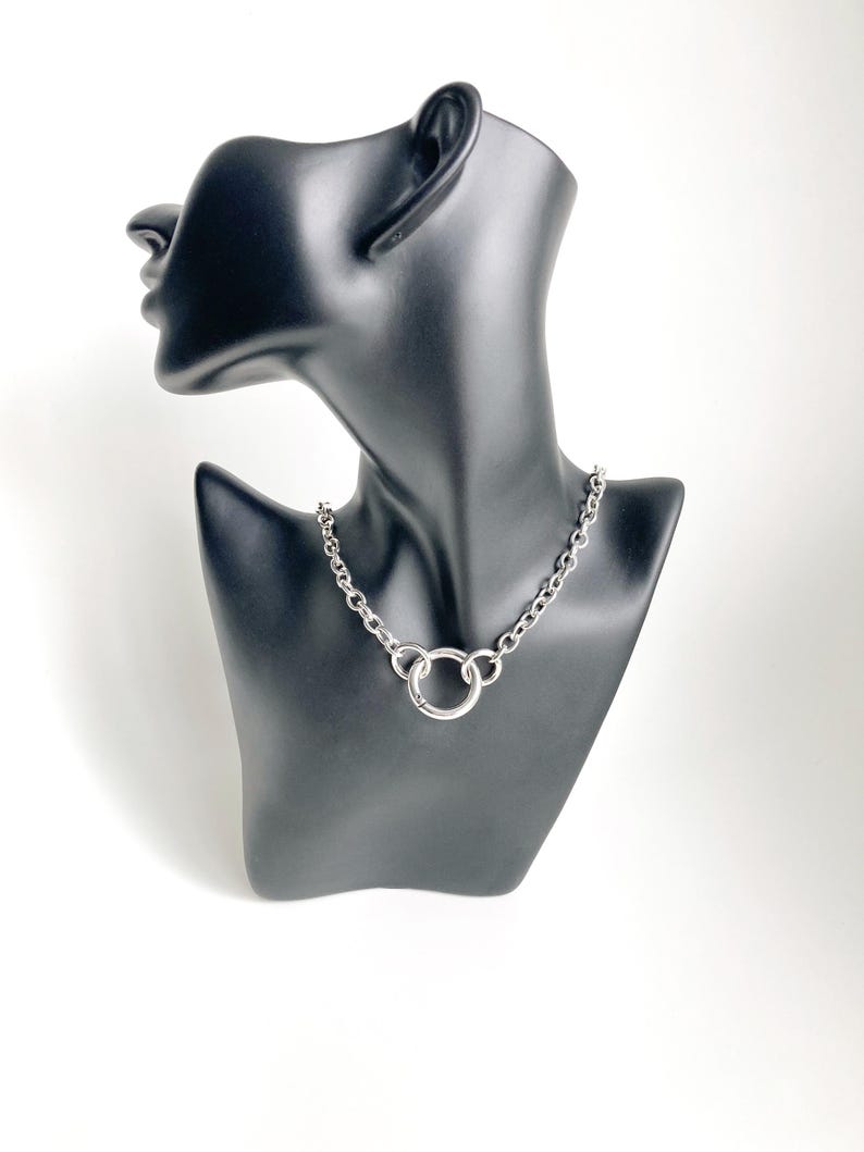 May include: A silver chain necklace with a large, round, silver pendant. The necklace is on a black mannequin.